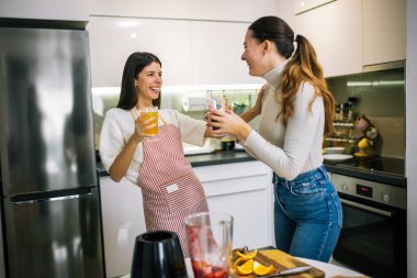 İki genç kadın modern bir mutfakta taze meyve suyu ve meyve sularının tadını çıkarıyorlar. Gülümsüyorlar ve sağlıklı, renkli bir içki anı paylaşıyorlar..