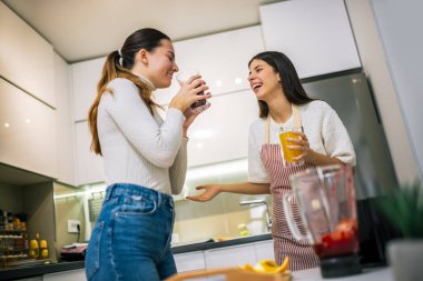 İki genç kadın modern bir mutfakta taze meyve suyu ve meyve sularının tadını çıkarıyorlar. Gülümsüyorlar ve sağlıklı, renkli bir içki anı paylaşıyorlar..