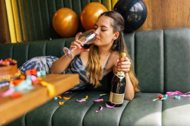 Yorgun bir kız, etrafı konfeti, pasta, hediye ve partilerle çevrili dağınık bir kanepede şarap içer.