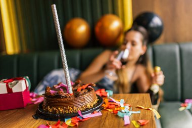 Yorgun bir kız, etrafı konfeti, pasta, hediye ve partilerle çevrili dağınık bir kanepede şarap içer.