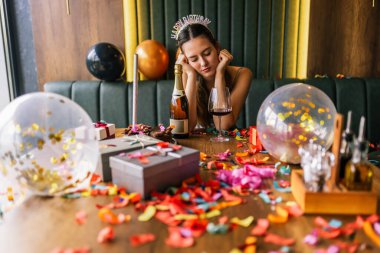 Yorgun bir kız, etrafı konfeti, pasta, hediye ve partilerle çevrili dağınık bir kanepede şarap içer.