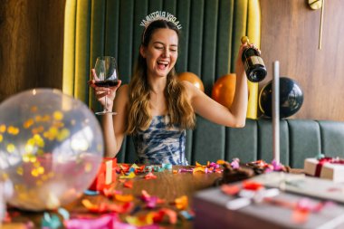 Yorgun bir kız, etrafı konfeti, pasta, hediye ve partilerle çevrili dağınık bir kanepede şarap içer.