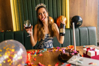 Yorgun bir kız, etrafı konfeti, pasta, hediye ve partilerle çevrili dağınık bir kanepede şarap içer.