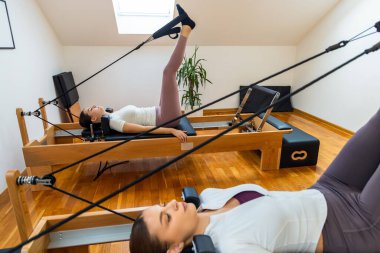 İki kadın sırt üstü yatar pilates reformcuları, kayışları yukarı doğru çekerler, çekirdekleri, bacakları birbirine bağlarlar ve esneklik ve gücü artırırlar..