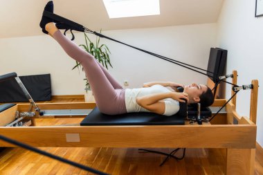 Bir kadın sırt üstü bir pilates reformcusunun üzerine uzanır, bacakları ile kayışları yukarı doğru çekerek çekirdek, bacak ve dayanıklılığı, esnekliği ve dengeyi arttırır..