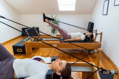 İki kadın sırt üstü yatar pilates reformcuları, kayışları yukarı doğru çekerler, çekirdekleri, bacakları birbirine bağlarlar ve esneklik ve gücü artırırlar..