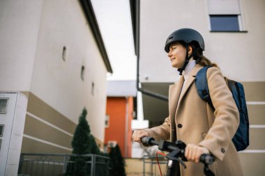 Şehirde elektrikli scooter kullanan genç bir kadın, kask ve sırt çantası takıyor, şehir hareketliliğini, bağımsızlığını ve aktif yaşam tarzını gösteriyor..