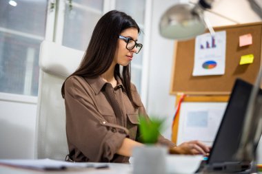 Modern bir ofiste dizüstü bilgisayarı üzerinde çalışan genç profesyonel bir kadın, görevlere odaklanmış ve kurumsal bir ortamda verimlilik gösteriyordu..