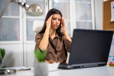 Modern bir ofiste masasında oturan genç profesyonel bir kadın, baş ağrısı yüzünden başını tutuyor, iş sırasında yorgunluk ve rahatsızlık gösteriyor..