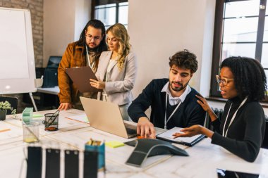 Siyahi bir kadın da dahil olmak üzere farklı ırktan genç iş adamları konferans odasında dizüstü bilgisayarda oturmuş, fikirleri tartışıyor ve işbirliği yapıyor, takım çalışması ve çeşitliliği vurguluyor..