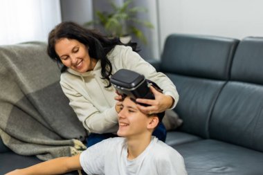 İlgili bir anne, genç oğluna modern bir oturma odasında sanal gerçeklik kulaklığı takmasında ya da ayarlamasında yardım ediyor..