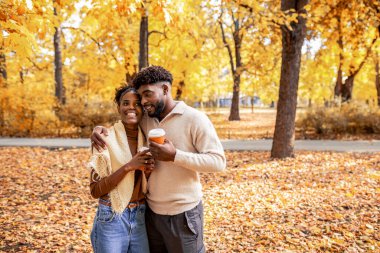 Afro-Amerikan çift, sonbahar parkında kahve fincanlarıyla yürüyor, konuşuyor ve gülümsüyor, aşk, bağlantı ve sonbahar atmosferiyle dolu sıcak, romantik bir anı paylaşıyorlar..