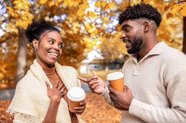 Afro-Amerikan çift, sonbahar parkında kahve fincanlarıyla yürüyor, konuşuyor ve gülümsüyor, aşk, bağlantı ve sonbahar atmosferiyle dolu sıcak, romantik bir anı paylaşıyorlar..