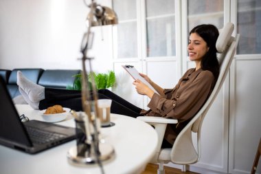 Genç bir kadın belgeleri incelerken ayaklarını masaya koyuyor. Konforlu ve rahat ofis sahnesi çoklu görev, üretkenlik ve modern yaşam tarzını gösteriyor.