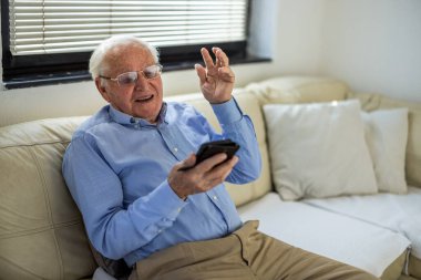 Evde oturan yaşlı bir adam cep telefonuyla video görüşmesi yapıyor. Yaşlılar için ideal yaşam tarzı, iletişim, teknoloji, uzak sağlık hizmetleri ve modern yaşam kavramları.