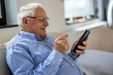 Evde otururken cep telefonu kullanan yaşlı bir adam.