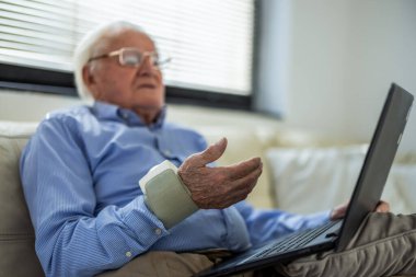 Evde yaşlı bir adam dizüstü bilgisayarla doktorla konuşurken kan basıncını ölçüyor. Teletıp, uzaktan sağlık, kıdemli bakım ve modern tıp teknolojisi kavramı.