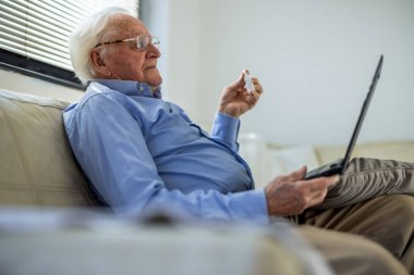 Evde yaşlı bir adam bilgisayardaki teletıp görüntülü konuşma sırasında doktora göz damlası gösteriyor. Uzaktan sağlık hizmetleri, kıdemli bakım, tıbbi danışma ve evde tedavi kavramları için ideal.