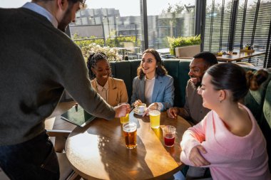 Garson, bir kafedeki masada oturan bir grup arkadaşa içecek getiriyor. Dostça bir hizmet veriyor, sosyalleşiyor ve neşeli bir atmosferde konukseverlik gösteriyor..