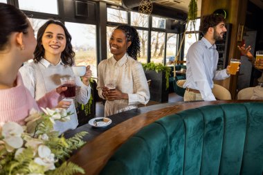 İki erkek bir arada duruyor ve üç kadın ayrı ayrı bir kafede, her biri kendi sohbetlerine dalmış, rahat bir ortam ve sosyalleşme sergiliyor..