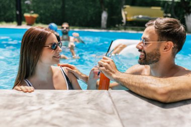 Güneş gözlüklü mutlu genç çift yüzme havuzunda kokteyllerin tadını çıkarıyor, birbirlerine bakıyor ve birlikte eğlenceli, rahatlatıcı bir yaz anını paylaşıyorlar..