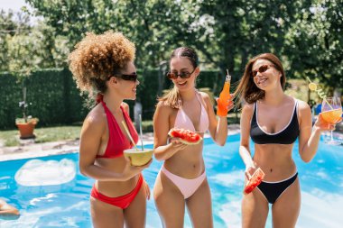 Yüzme havuzunda rahatlayan, kokteyl içen ve taze karpuz yiyen, yaz havasının ve arkadaşlığın tadını çıkaran bir grup çok kültürlü kadın..