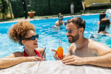 Güneş gözlüklü mutlu genç çift yüzme havuzunda kokteyllerin tadını çıkarıyor, birbirlerine bakıyor ve birlikte eğlenceli, rahatlatıcı bir yaz anını paylaşıyorlar..