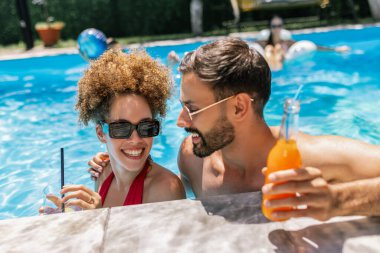 Güneş gözlüklü mutlu genç çift yüzme havuzunda kokteyllerin tadını çıkarıyor, birbirlerine bakıyor ve birlikte eğlenceli, rahatlatıcı bir yaz anını paylaşıyorlar..