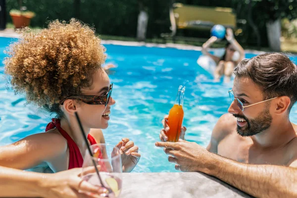 Güneş gözlüklü mutlu genç çift yüzme havuzunda kokteyllerin tadını çıkarıyor, birbirlerine bakıyor ve birlikte eğlenceli, rahatlatıcı bir yaz anını paylaşıyorlar..