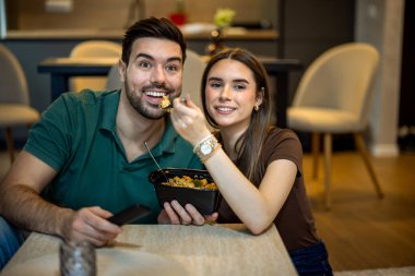 Genç bir çift yerde oturmuş makarna yiyor ve oturma odalarında televizyon izliyor..