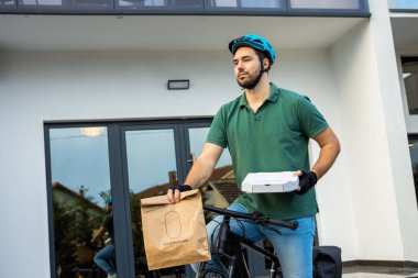 Kurye, elinde bir pizza kutusu ve bir kese kağıdıyla binanın önünde bisikletle dikilirken..