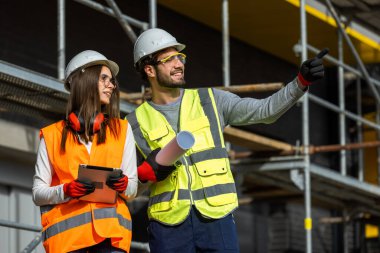 Genç erkek ve kadın mühendisler bir inşaat alanındaki dijital tablet ve kağıt planlarını inceliyorlar..