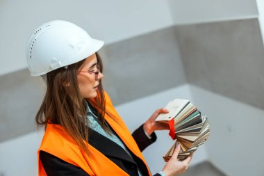 Beyaz güvenlik kaskı ve turuncu yelek giyen profesyonel bir kadın mimar banyoda renk paleti tutuyor. AP 'nin iç dekorasyonu için en iyi tasarımı seçmek için duvardaki farklı fayans gölgelerini dikkatlice karşılaştırıyor.