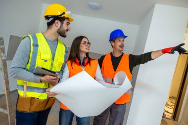 Güvenlik kaskı ve yüksek görüş açısına sahip yelekler giyen üç inşaat uzmanından oluşan profesyonel bir grup, parlak ve boş bir dairenin merkezinde toplandı. Ayrıntılara dikkat çekerek ve yenilemeyi tartışarak büyük bir kağıt planı tutuyorlar.