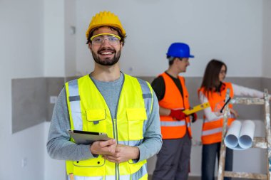 30 'lu yaşlarda kendine güvenen bir inşaat işçisinin profesyonel portresi, güvenlik kaskı ve parlak turuncu yüksek görüş mesafesi olan bir yelek giyiyor. Dostane bir gülümseme ile doğrudan kameraya bakıyor, elinde modern bir dijital tablet tutuyor..