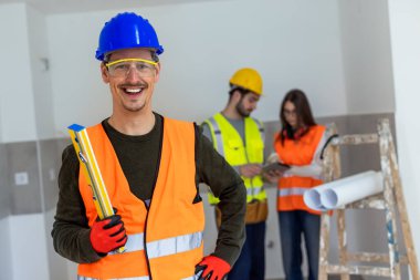Ruhani bir seviyede tutarken kameraya gülümseyen bir inşaat işçisinin profesyonel portresi. Güvenlik kaskı ve yüksek görüş mesafesi olan bir yelek giyiyor..