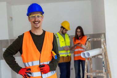 Elinde ölçüm cetveli tutarken kameraya gülümseyen bir erkek inşaat işçisinin profesyonel portresi. Güvenlik kaskı ve yüksek görüş mesafesi olan bir yelek giyiyor..