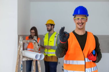 Bir erkek inşaat işçisinin neşeli ve cesaret verici bir portresi. Geniş bir gülümsemeyle doğrudan kameraya bakıyor. Başparmak işareti yapıyor..
