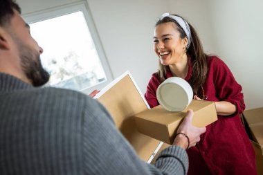 Geniş, parlak ve boş bir dairede duran bir adam ve kadın. Her biri başarılı bir taşınma gününü simgeleyen karton bir kutu taşıyor..