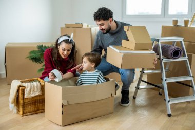 Genç bir aile taşınma gününde, bir anne çocuğuyla karton bir kutunun içinde oynarken baba arka plandaki taşınmayı yönetir.