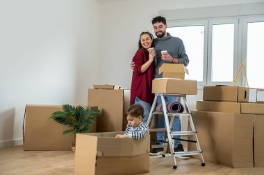 Parlak, boş bir dairede hareket halindeki bir kutunun içinde oturan genç bir adam ve bir kadın birlikte durup çocuklarına gülümsüyor..