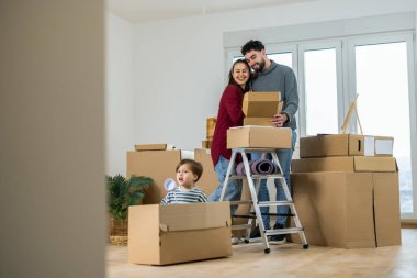Parlak, boş bir dairede hareket halindeki bir kutunun içinde oturan ve çocuklarına gülümseyen genç bir adam ve kadın..