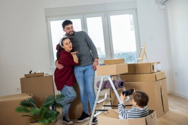 Küçük bir çocuğun karton bir kutuda oturduğu ve anne ve babasının fotoğrafını çekmek için akıllı telefon kullandığı yeni bir dairede eğlenceli bir sahne..