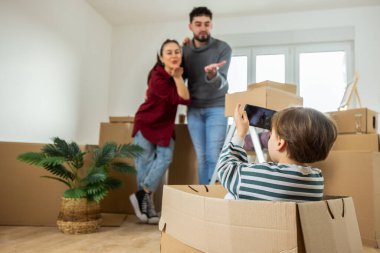 Küçük bir çocuğun karton bir kutuda oturduğu ve anne ve babasının fotoğrafını çekmek için akıllı telefon kullandığı yeni bir dairede eğlenceli bir sahne..