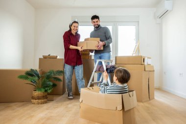 Taşınırken küçük bir çocuğun güvenli bir şekilde karton bir kutuda oturduğu hareketli bir aile sahnesi, anne ve babasının fotoğrafını çekmek için akıllı telefon kullanıyor. Ebeveynler boş oturma odasında büyük kutular taşıyarak çalışmakla meşgul.