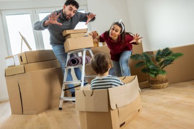 Taşındıkları gün, genç bir erkek ve kadının yürümeye başlayan çocuklarına aptal suratlar yapmak için karton bir kutunun üzerine eğildikleri neşeli ve samimi bir an. Kutunun içinde oturan çocuk neşeyle gülüyor..