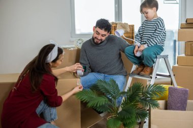 Genç bir çift ve küçük çocukları kutuları taşımak için birlikte çalışıyorlar. Ebeveynler yerde diz çökmüş, ellerinde bir rulo bant tutarken, bebek bakınıyor..