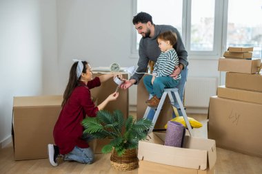 Genç bir çift ve küçük çocukları kutuları taşımak için birlikte çalışıyorlar. Ebeveynler yerde diz çökmüş, ellerinde bir rulo bant tutarken, bebek bakınıyor..