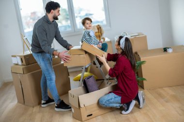 Genç bir anne, baba ve küçük oğulları parlak, boş oturma odalarında kutuları taşıyorlar. Ebeveynler daha büyük kutular taşırken, küçük çocuk gururla anne ve babasını taklit ederek küçük bir kutu taşıyor..