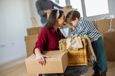 Yeni bir dairede, bir annenin bebeğiyle oynamak için diz çöktüğü ve onu güldürdüğü şefkatli bir aile anı. Bulanık arkaplanda baba kutuları düzenleyip açmakla meşgul görünüyor..